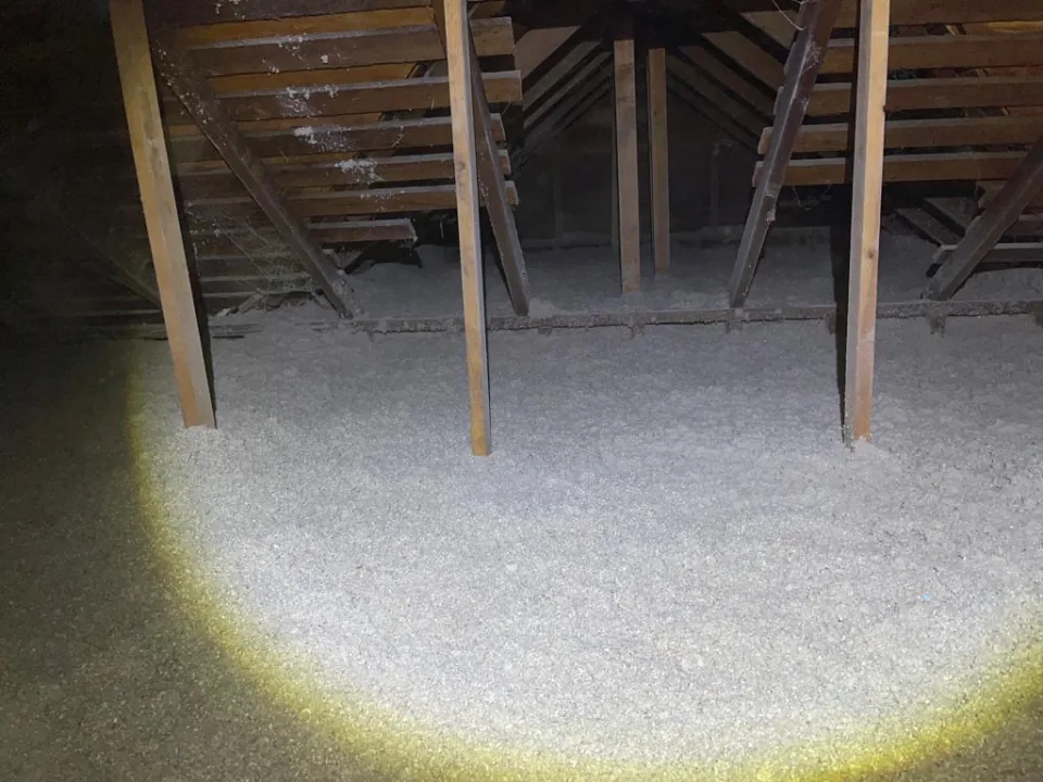 The same attic after the job, lit by a flashlight: an even layer of fresh blown-in cellulose across the floor between the posts, the gable end behind
