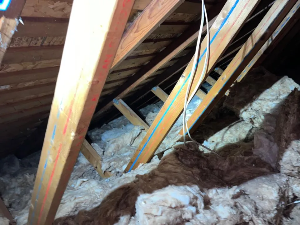 Attic before the job: torn old brown and white batt insulation piled unevenly between marked roof trusses, a loose cable hanging down